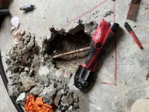 Concrete floor with a hole revealing a copper pipe, surrounded by debris and tools, including a red power tool and a red marker. Cowboyplumbingtx ensures precision in every step of the repair process.