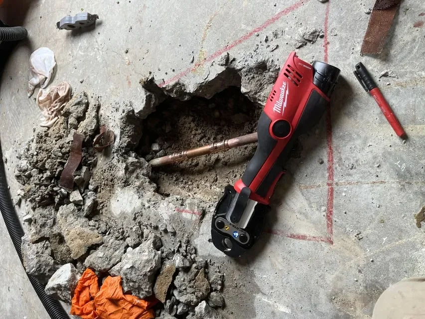 Concrete floor with a hole revealing a copper pipe, surrounded by debris and tools, including a red power tool and a red marker. Cowboyplumbingtx ensures precision in every step of the repair process.