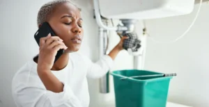 A woman with short, light-colored hair is on the phone, appearing concerned as she stands in front of a sink. She holds a cleaning cloth in one hand and is positioned next to a green bucket. The background is bright and minimalistic, emphasizing her focused expression while dealing with a plumbing issue.