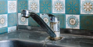 The image shows a kitchen faucet mounted on a sink, with visible signs of rust and mineral buildup around its base, suggesting wear and neglect. The background features decorative blue and white patterned tiles with intricate, symmetrical designs, adding a vintage or traditional aesthetic to the setting.