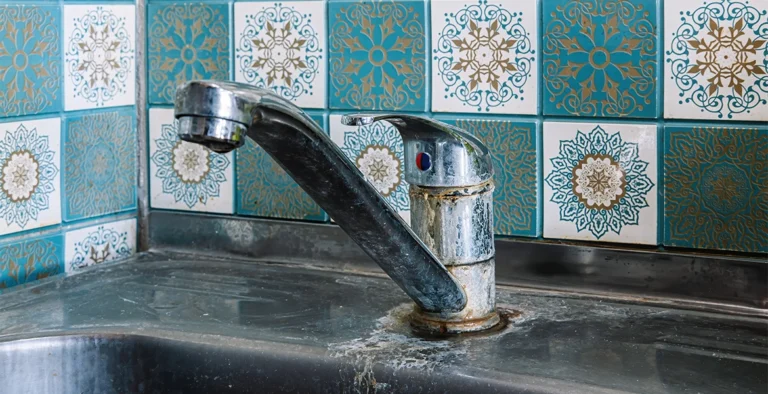 The image shows a kitchen faucet mounted on a sink, with visible signs of rust and mineral buildup around its base, suggesting wear and neglect. The background features decorative blue and white patterned tiles with intricate, symmetrical designs, adding a vintage or traditional aesthetic to the setting.